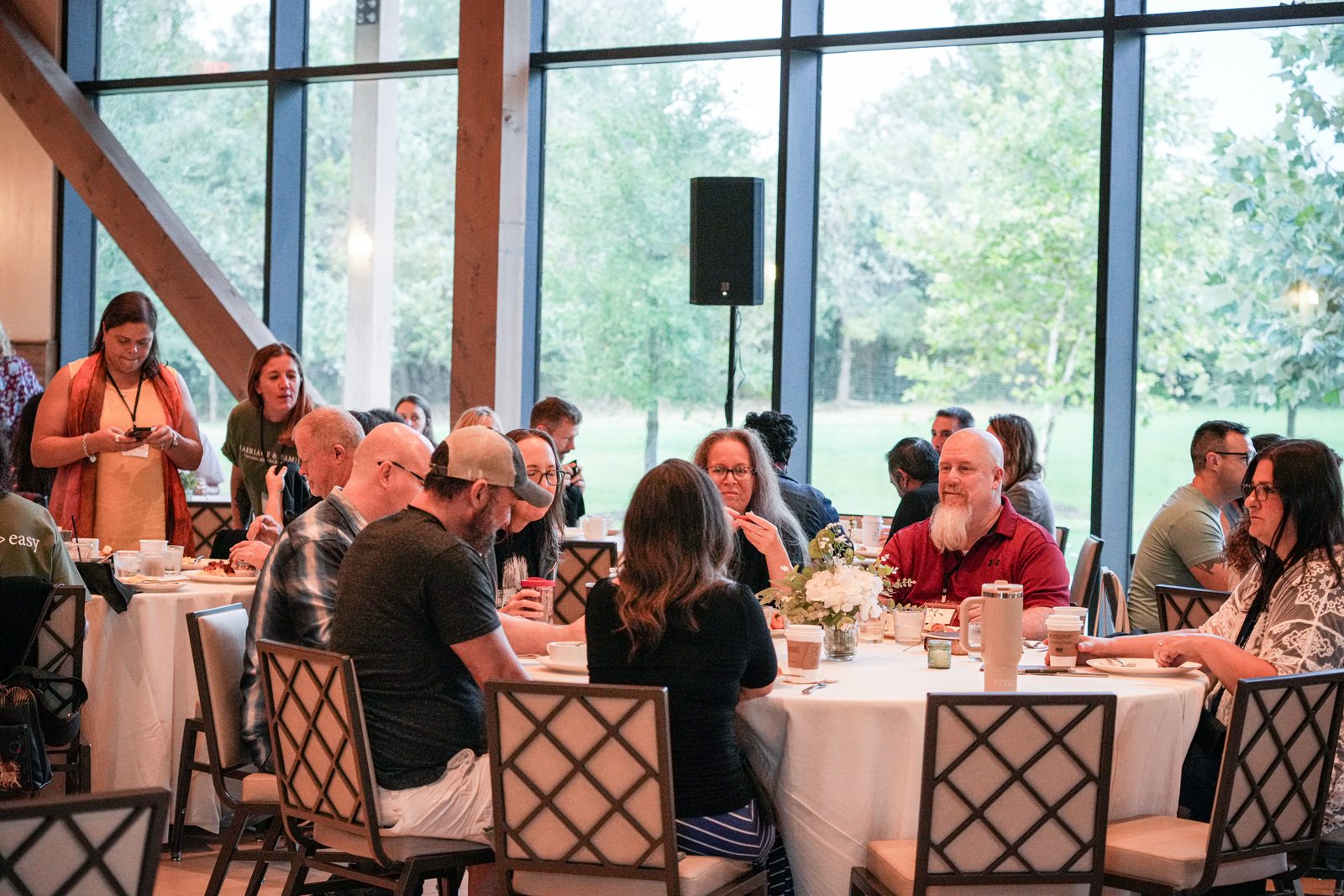 Couples at breakfast around a round table in the Riverbend Barn, trees showing through the glass wall behind them
