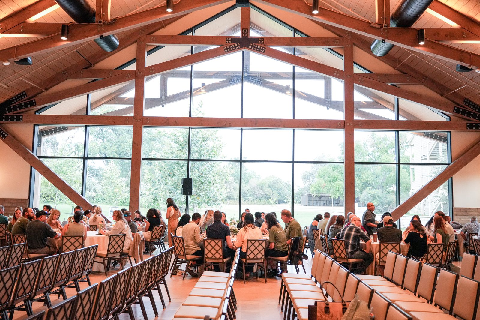 The Riverbend Barn at Hyatt Regency Lost Pines full of couples at round tables under the timber trusses and the tall glass wall